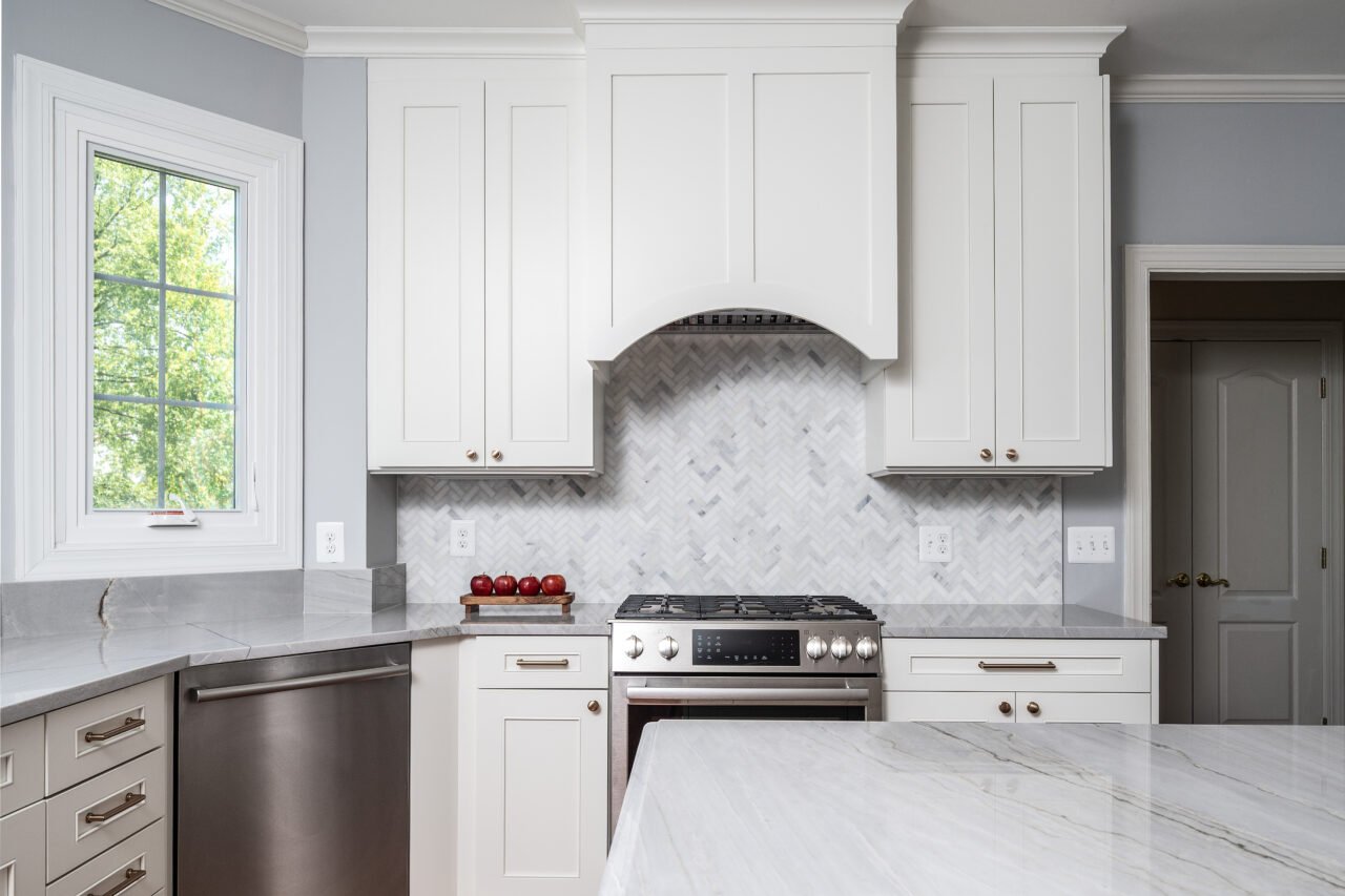 Custom kitchen remodel in Leesburg, VA with white shaker cabinetry, decorative arched range hood, herringbone tile backsplash, and stainless steel gas range.