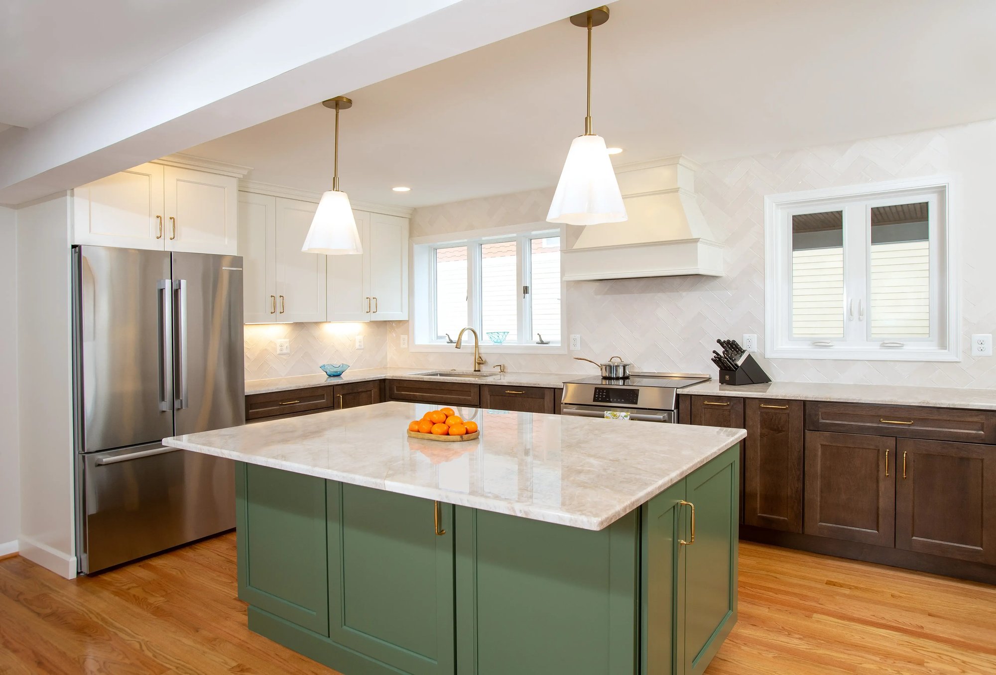 Angled view of a contemporary kitchen featuring large center island with green cabinets and brass fixtures in front of a refrigerator and stove underneath a custom white range hood