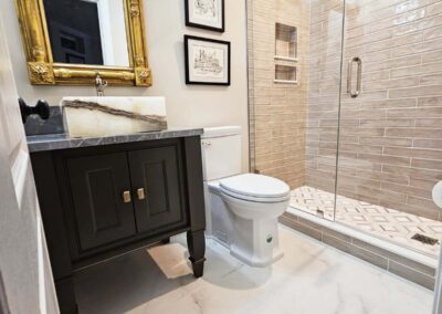 Elegant bathroom featuring a black vanity with a gold-framed mirror, a modern toilet, and a glass-enclosed shower with beige subway tiles and geometric patterned floor tiles.